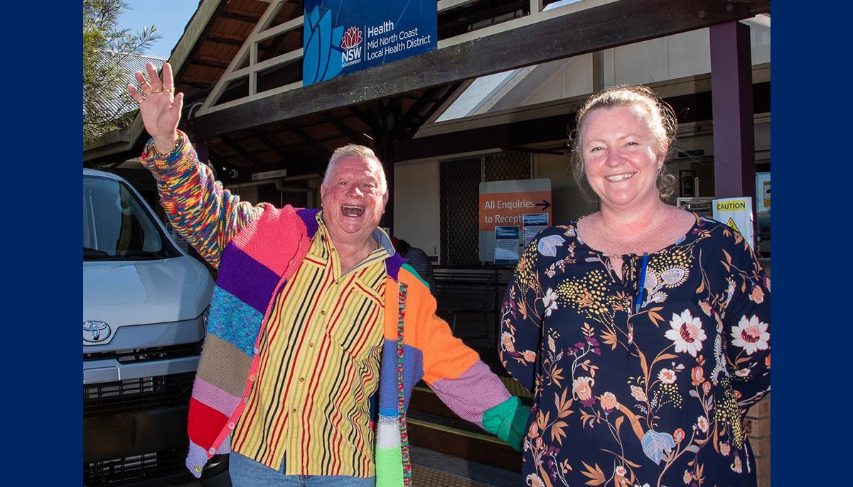 man wearing very colourful cardigan with hand raised in the air and a big, open smile on his face standing next to a nurse outside a hospital.