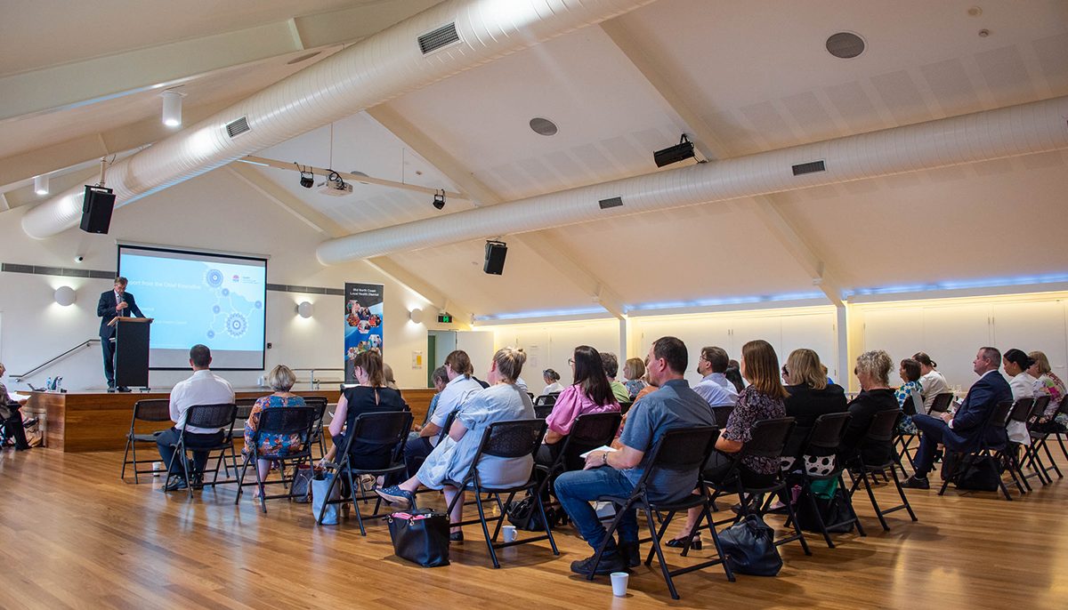 people seated in a large room looking forward to a stage where a man in a suit is speaking at a lecturn.