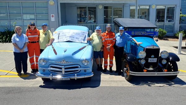 A group of people standing with two restored cars outside a hospital.