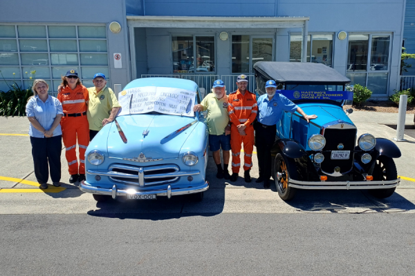 A group of people standing with two restored cars outside a hospital.