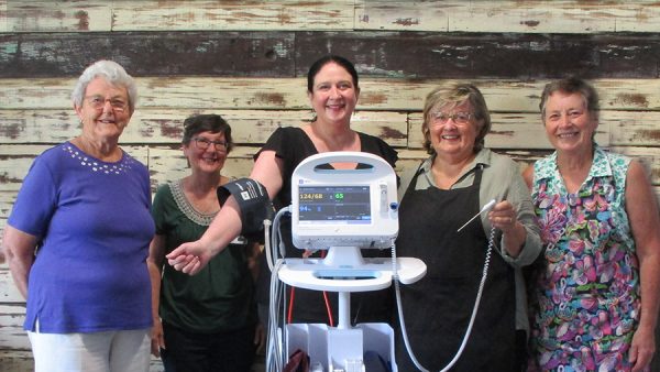 Five women standing against a timber wall. In front of them is a vital signs monitor.