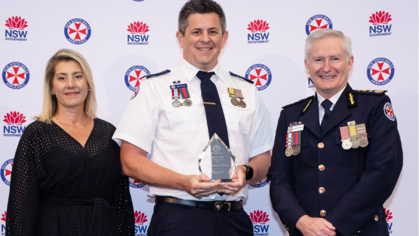 Three people smiling. The person in the centre is holding a trophy.