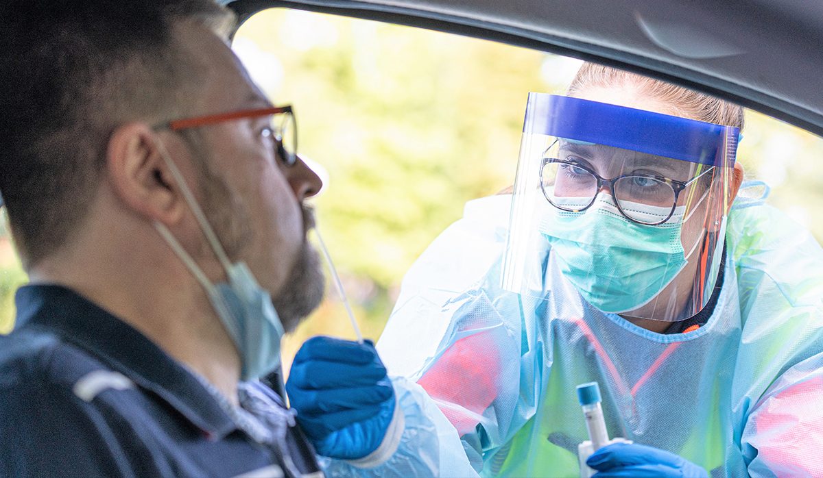 male driver in stationary car being tested for COVID by nurse wearing personal protective equipment