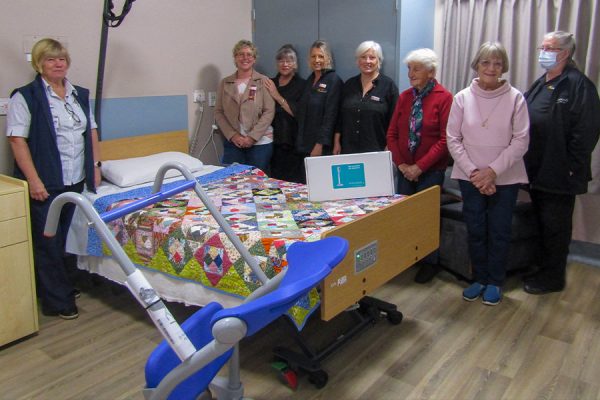 eight women, including a nurse, standing around a hospital bed.