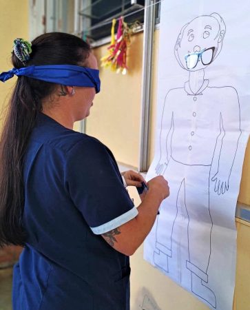 An enrolled nurse blindfolded pinning cardboard glasses on a paper sheet featuring the outline of a man.