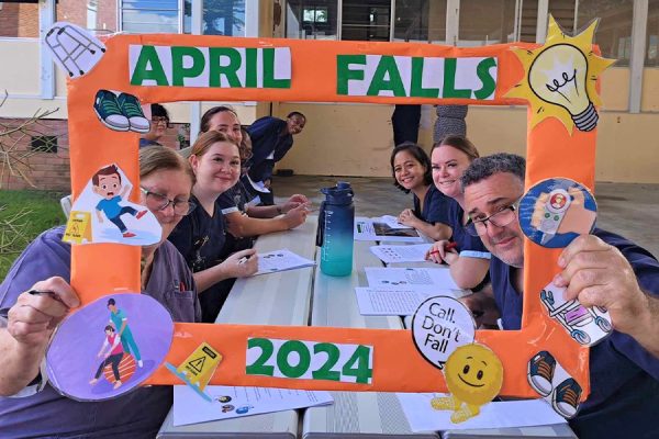 Nine nurses behind a cardboard picture frame featuring the word April Falls 2024.