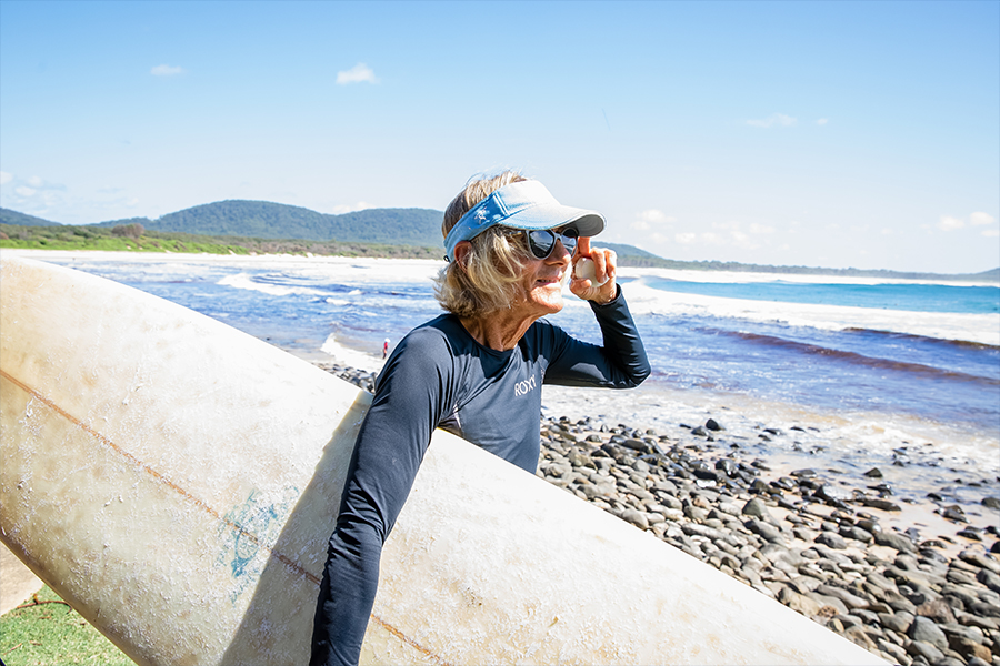 Woman wearing a hat and sunglasses, holding a surfboard and looking out towards the ocean.