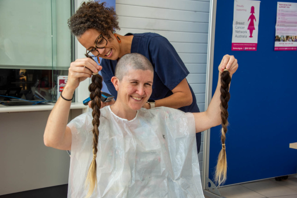 A woman shaving the hair of another woman who is holding up two plaits.