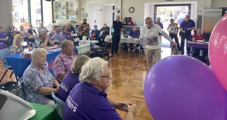 A room with people seated, many wearing purple or holding purple balloons.
