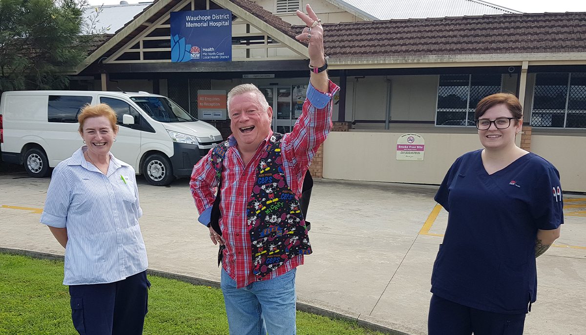 A man wearing colourful clothes standing between two nurses outside a hospital.