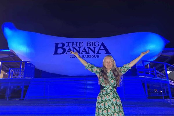 A young woman standing with arms outstretched in from of the Big Banana at Coffs Harbour, lit blue for lymphoedema week.