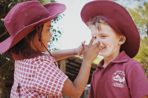 Two school children. One applying sunscreen to the face of the other.