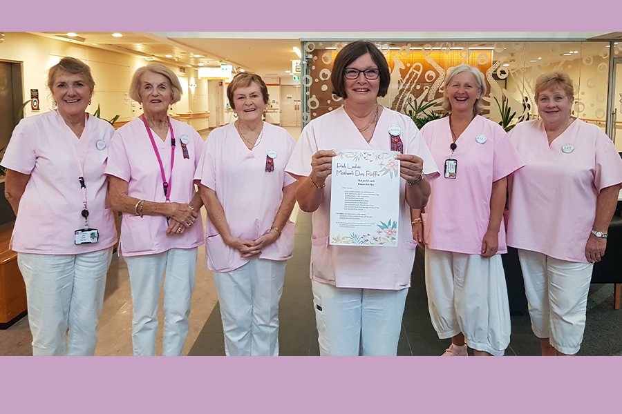 six ladies wearing pink smocks and white trousers standing together smily broadly while the one in the middle, a step fporward of the others, holds a flyer promoting a Mother's Day Raffle.