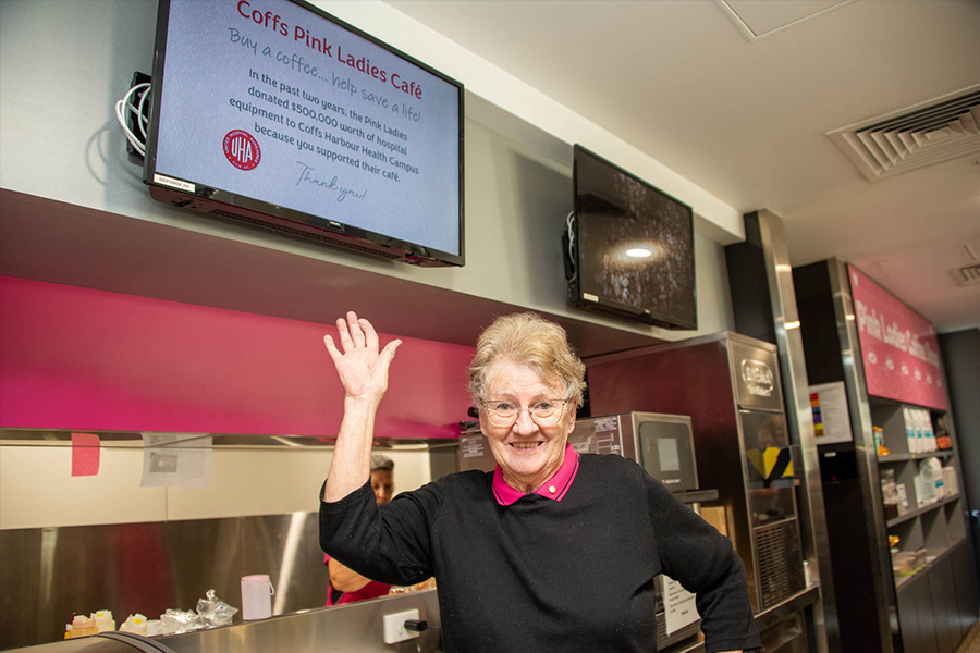 woman wearing a pink shirt and black jumper standing in a cafe with one arm in the air and a tv monitor behind her