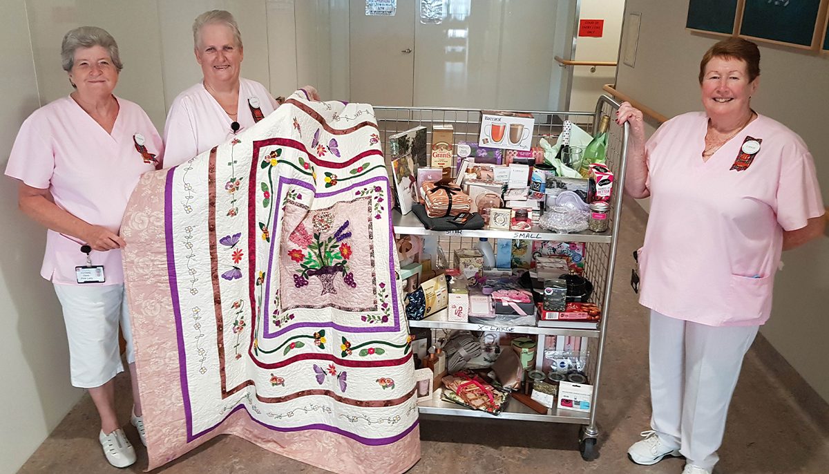 three women wearing pink smocks standing next to a trolley of raffle prizes.