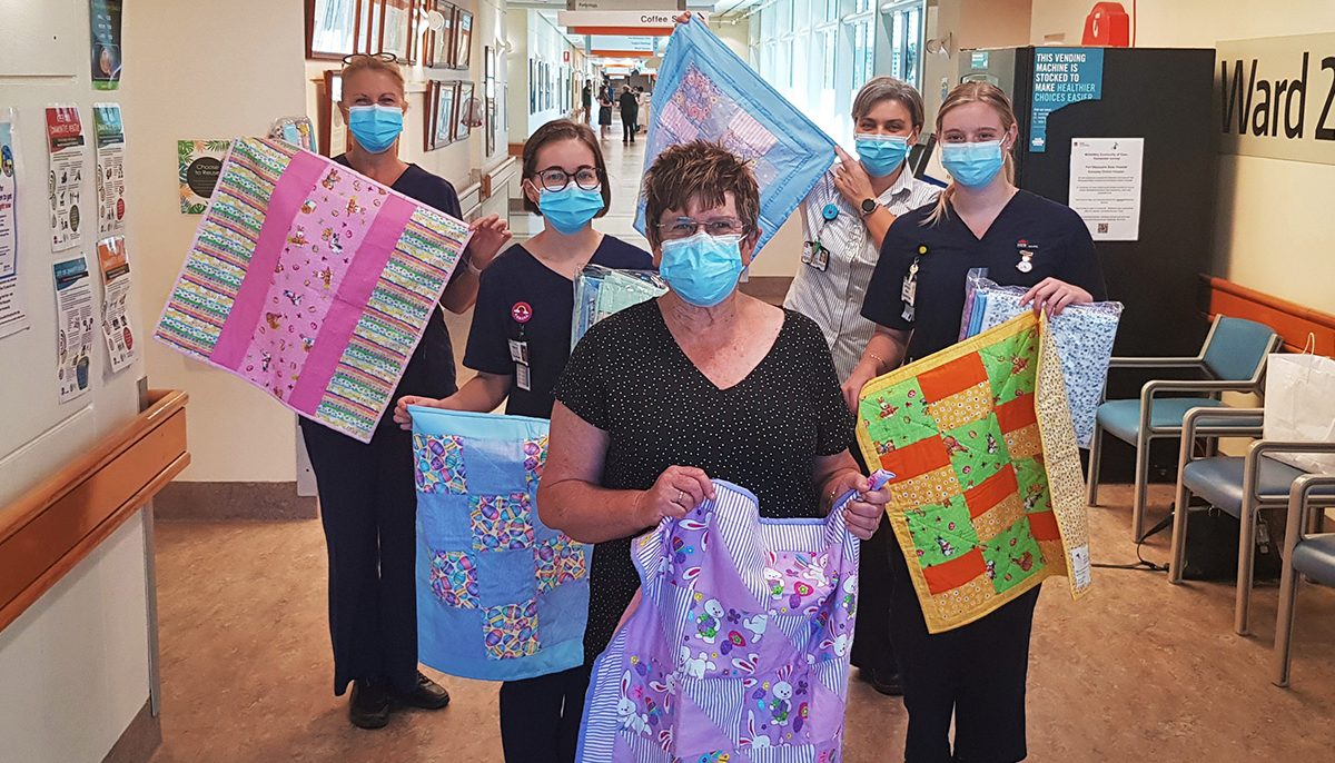 an older woman standing with midwives in a hospital corridor. All are holding quilts.