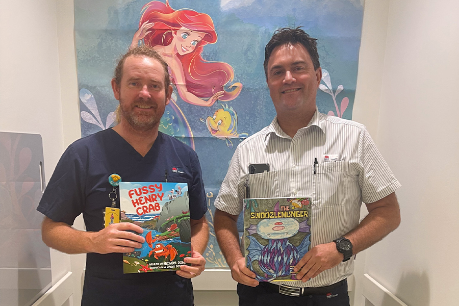 two men holding children's books. Both are nurses and behind them is a mural of Ariel, the Little Mermaid.
