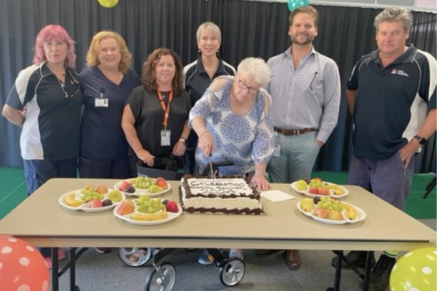 A group of healthcare workers with a senior citizen who is cutting a cake on a table with fruit platters.