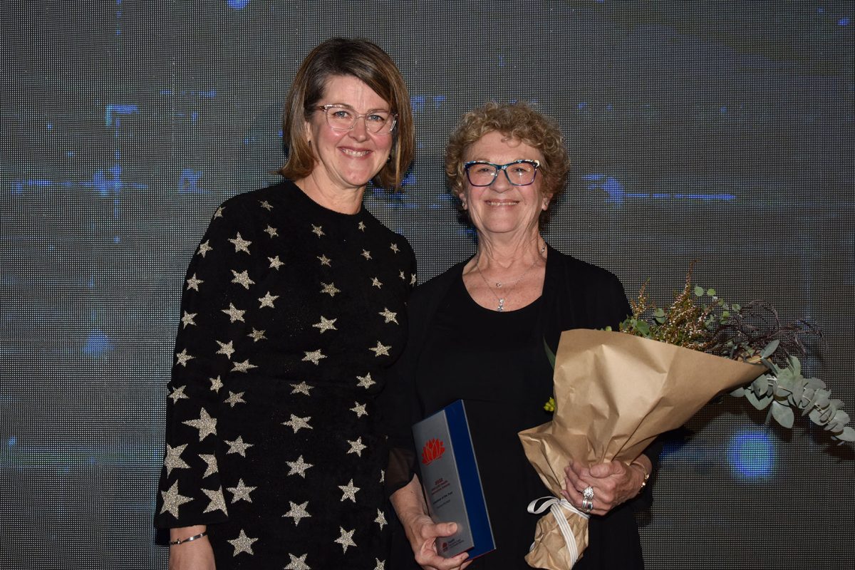 two women standing together - one holding an award and flowers