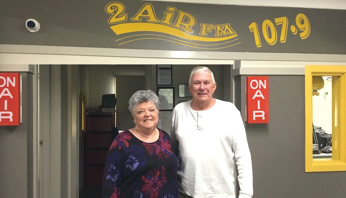 A woman and man standing under a sign in a radio studio which reads 2AIR FM 107.9.