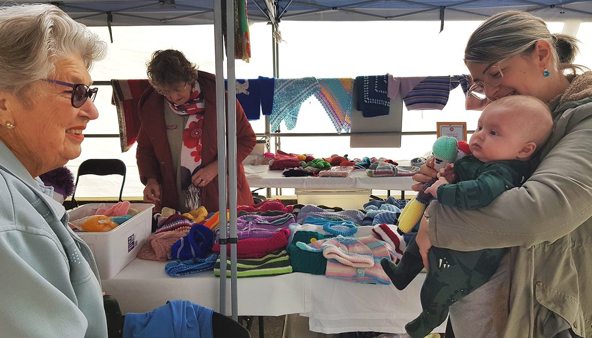 Older woman talking to a mum and her baby at a stall selling knitwear