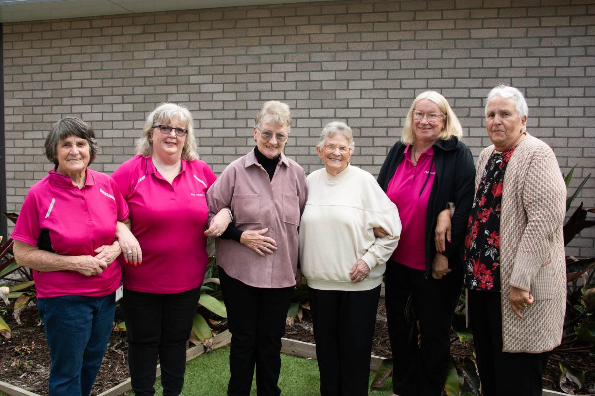 Six women standing side by side, some of the women are wearing pink shirts.