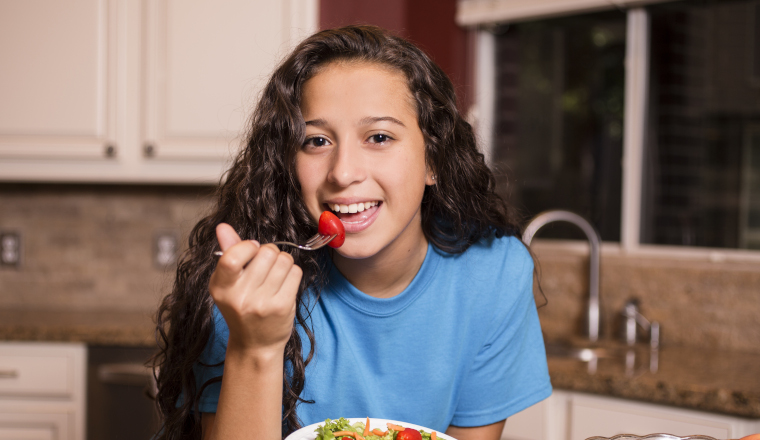 A teenage girl eats healthy salad after her school soccer team practice. She has a plate full of lettuce, carrots and tomatoes and also has a glass of water