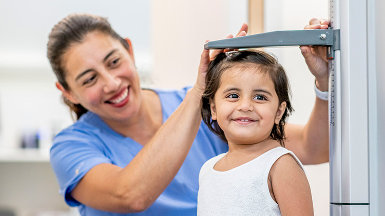 A nurse measures the height of a primary preschool-aged girl. They're both smiling.