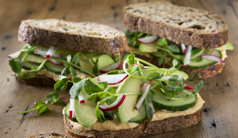 Three healthy vegetarian sandwich's on a rustic wood background. Sandwich's are on multigrain bread with roasted red pepper hummus , radish's ,cucumber , pea shoots and avocado.